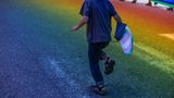 Kid holding trans pride flag walking down a rainbow-lit street